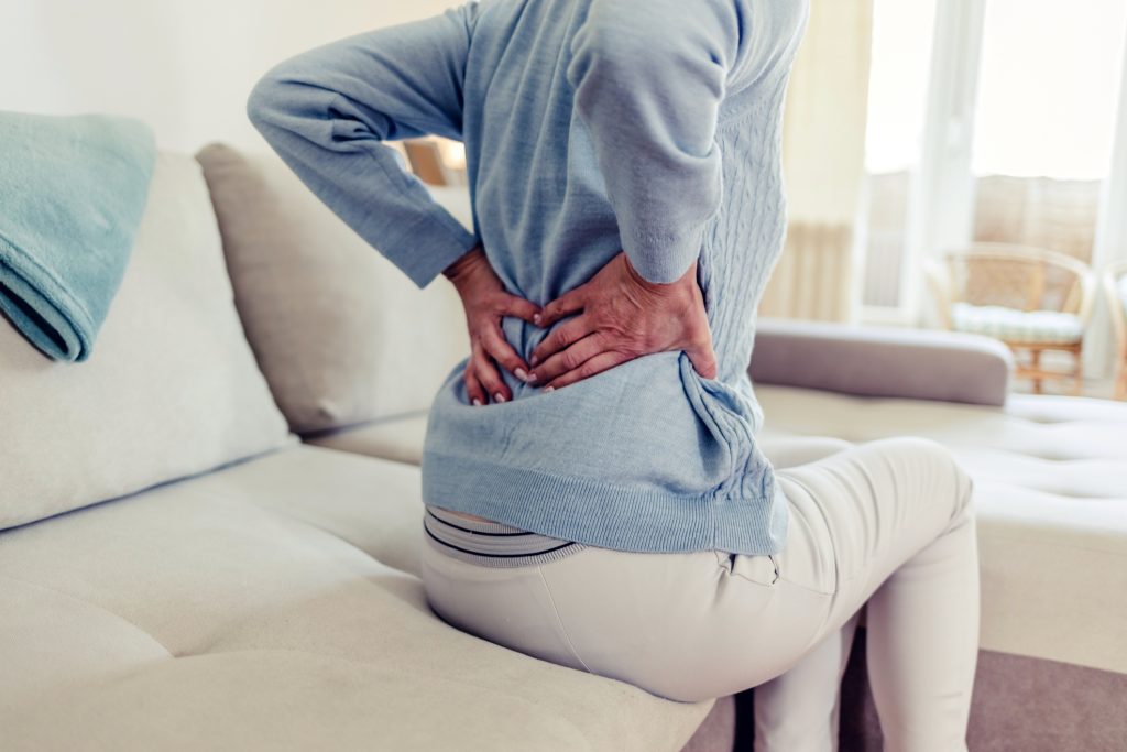 Cropped image of senior woman suffering from backache in morning sitting on bed, free space. Lower back pain in elderly person.